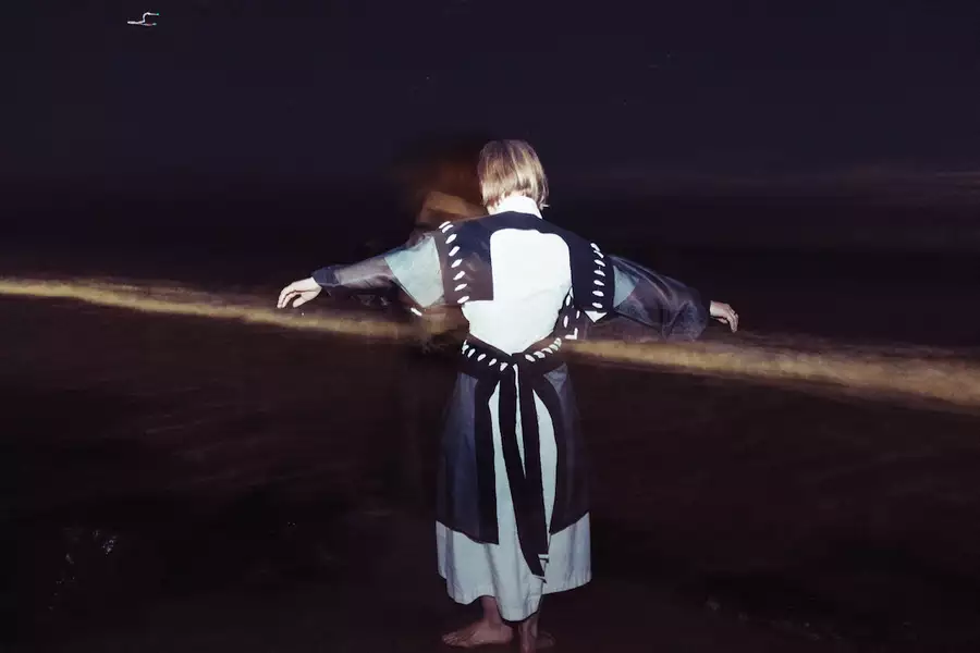 Amber Cronin stands barefoot at night with arms outstretched, wearing a robe. A long exposure effect creates a ghostly double image and a streak of light across the dark, shoreline background.