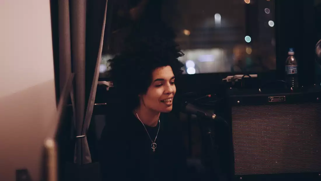 A woman sits inside, in front of a window with night outside, as she sings with a microphone in front of her.