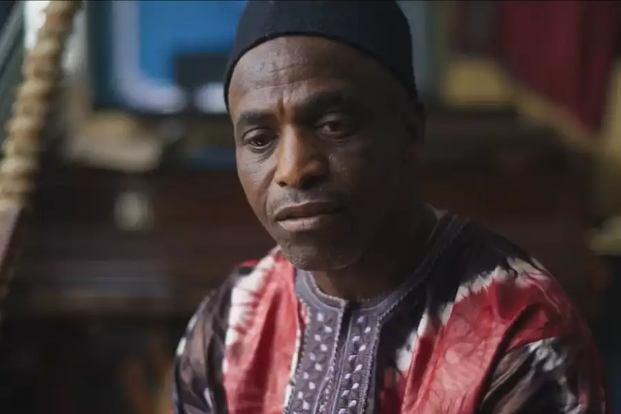 A man in traditional clothing sits indoors, looking thoughtfully at the camera with a stringed instrument beside him.