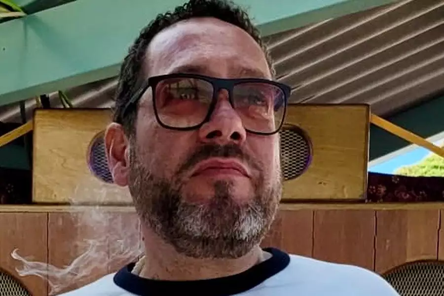 Dom stands outside in front of wooden speakers, wearing a white shirt with a dark collar. Smoke drifts beside his face, and a green roof structure is visible above.