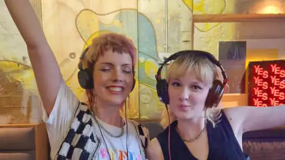 Two hosts of Is This It Radio show with headphones on. One has their hands in the air.