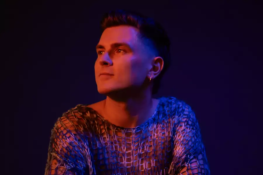 Fastlove wearing a meshed colourful top, looking to the side. The lighting is red.