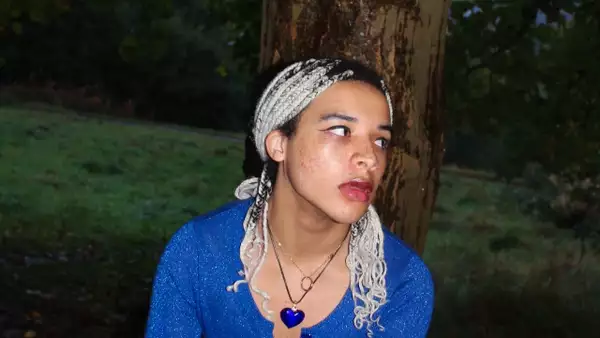 Lucas Assagba looking to the side, wearing a bright blue cardigan and heart-shaped necklace. Behind them is a forest.