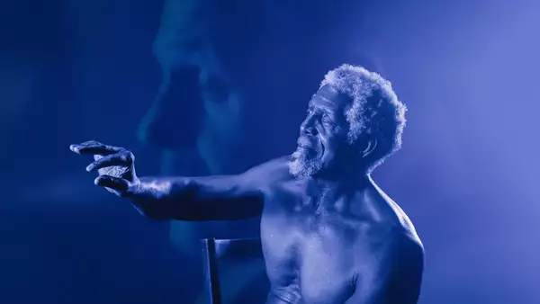 A shirtless elderly man with curly white hair and a beard sits on a chair, reaching out with an expressive hand. The image has a blue monochromatic tone with a soft-focus overlay of his face in the background.