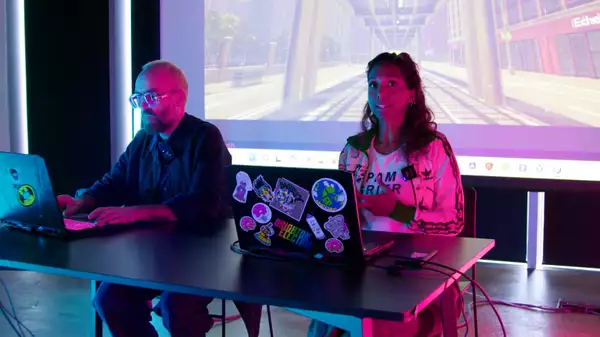 Simon Wilkinson and Myra Appannah sat at a desk on their computers. Behind them is a big screen showing a game.