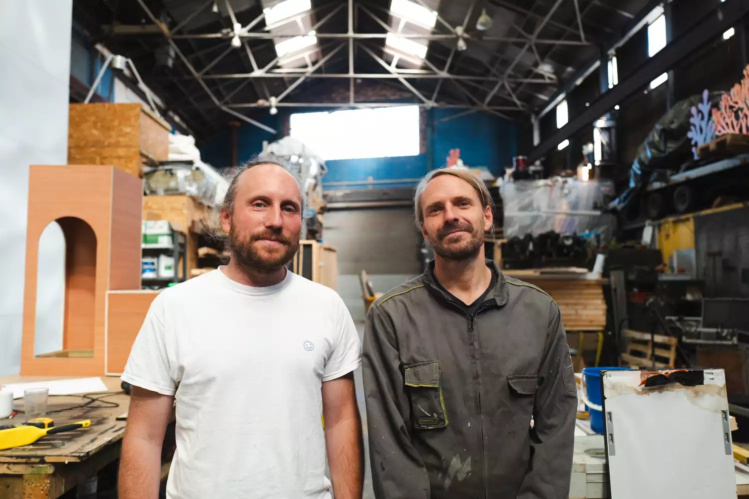 Two members of Studio Tonto stood in a Warehouse filled with equipment. One is wearing a white t-shirt and the other is wearing a grey jacket.