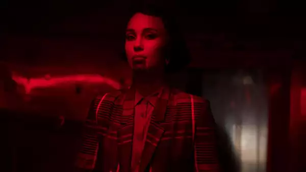 Kaneza Schaal in a dimly lit room bathed in intense red light, wearing a patterned jacket. The dramatic lighting and shadows create a cinematic atmosphere.