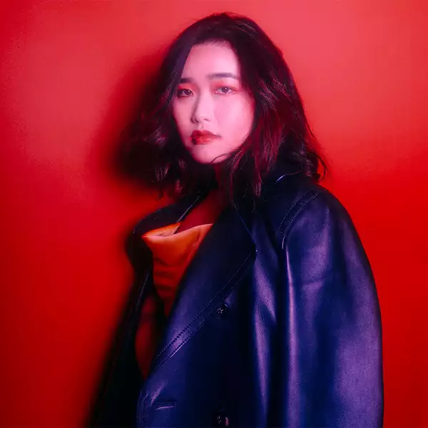 Edane stood against a red background. She is looking into the camera and holding a leather jacket draped over her shoulder