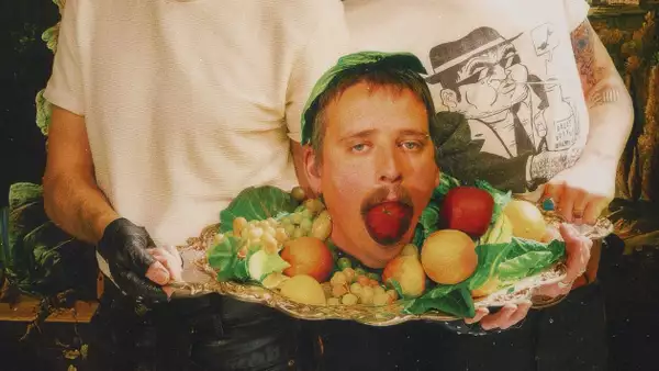 Two people hold a silver platter with a man’s head surrounded by fruit and leaves; the man’s mouth is open with a strawberry inside.