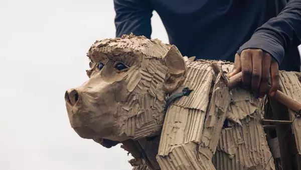 Close-up of a cardboard baboon puppet being operated by a person using wooden controls.
