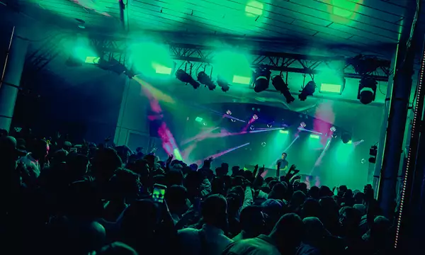 A DJ set on Festival Square from MIF23, showing a large crowd of people dancing and filming a DJ on stage. The lighting is hazy green and there are purple strobe lights.