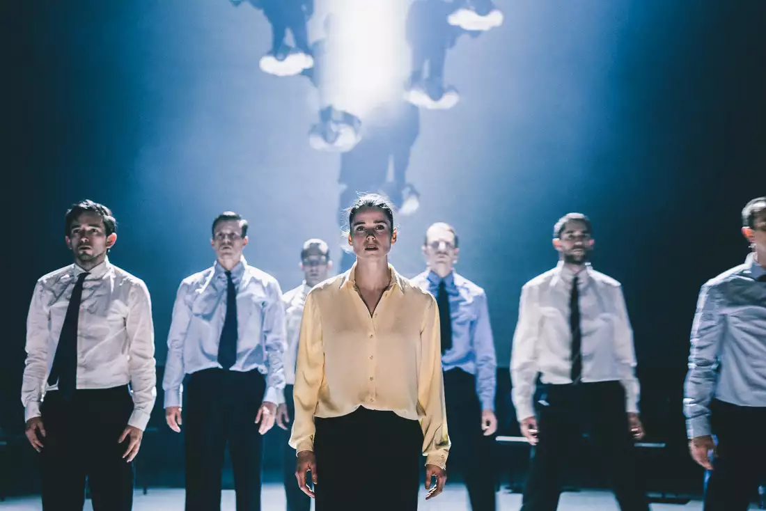 A still from Figures in Extinction [2.0] showing seven performers stood upright, wearing suits and staring at the audience