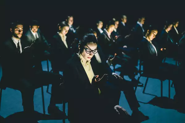 A still from Figures in Extinction [2.0] showing performers sat on chairs, wearing glasses and suits and looking at their phones