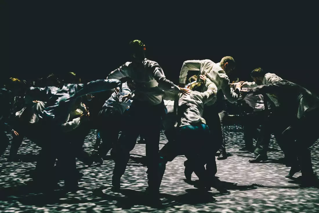 A still from Figures in Extinction [2.0] showing performers on stage wearing suits, making large contorted shapes with their bodies under spotted lighting