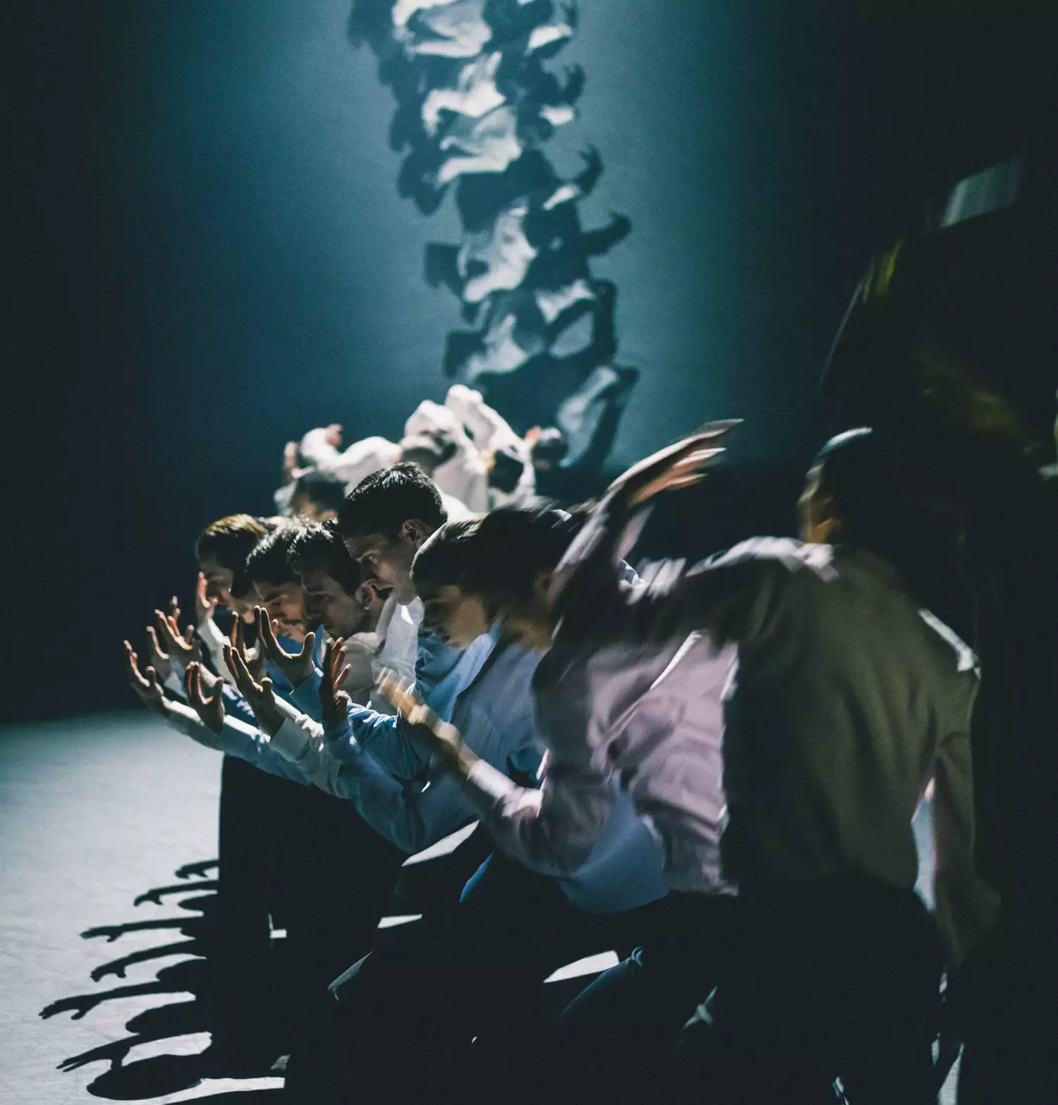 Dancers in a row wearing button up shirts reaching upwards with an overhead shot of their movements projected onto a screen behind
