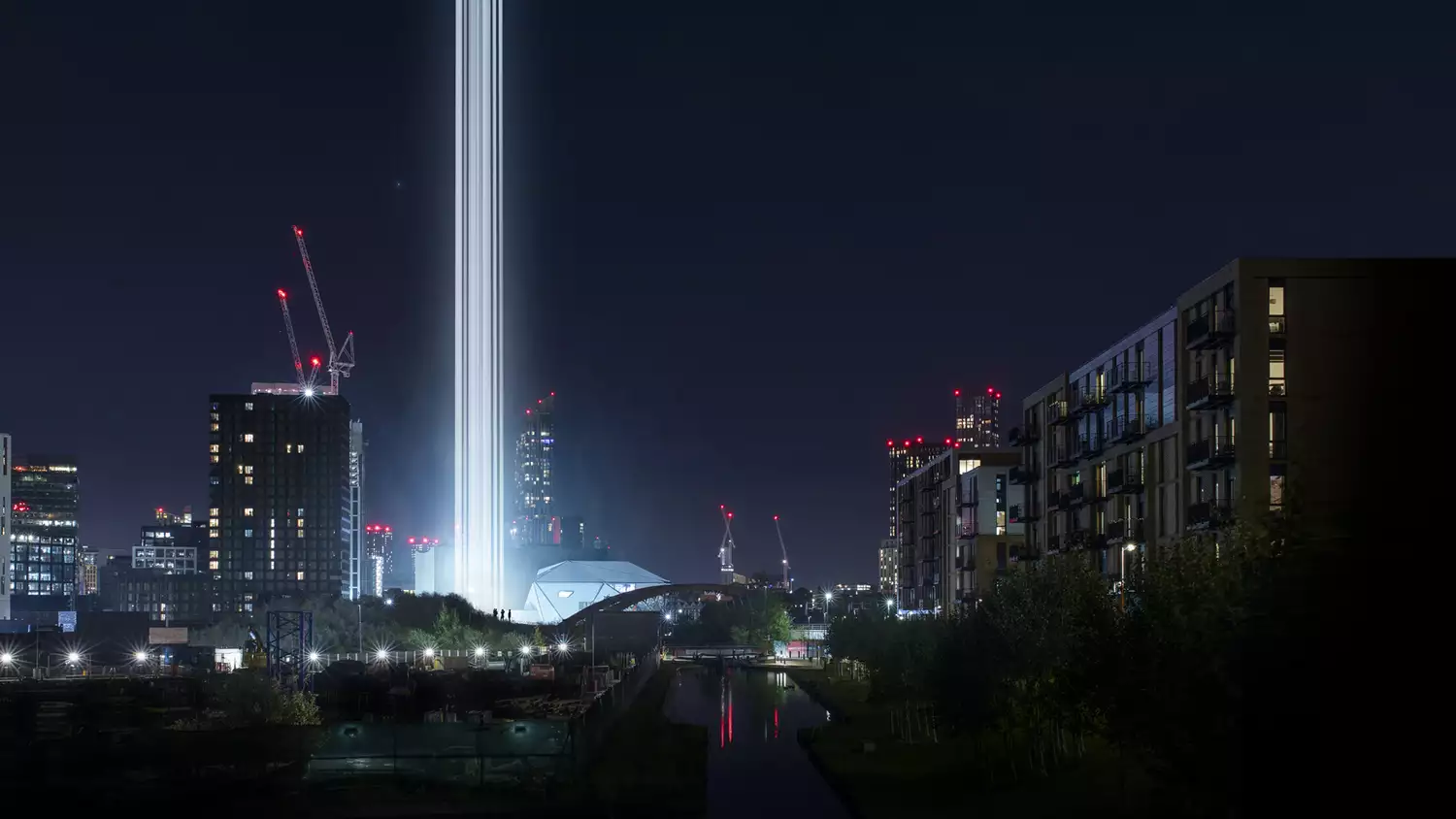 A Manchester cityscape at nighttime with bright columns of light visible next to the Factory International building.