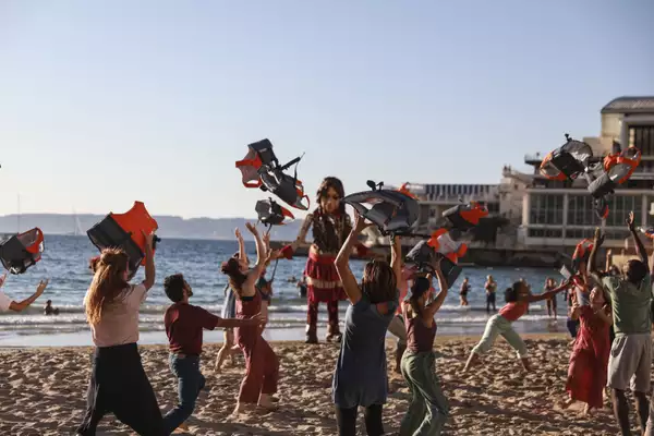 Performers on a sunny Marseille beach throw orange and black life jackets into the air. In the background, the Little Amal puppet stands in front of the sea