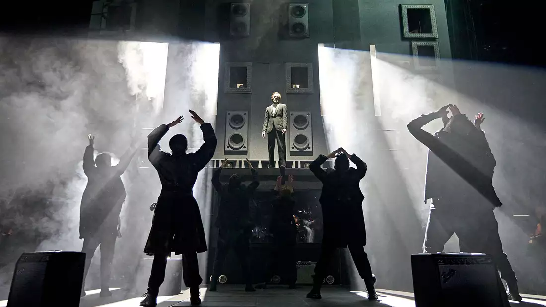 Production shot from Hamlet Hail to the Thief, which shows Claudius stood on a large structure. The cast are turned towards him and the background is smoky.