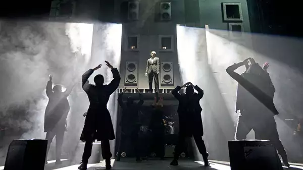 Production shot from Hamlet Hail to the Thief, which shows Claudius stood on a large structure. The cast are turned towards him and the background is smoky.