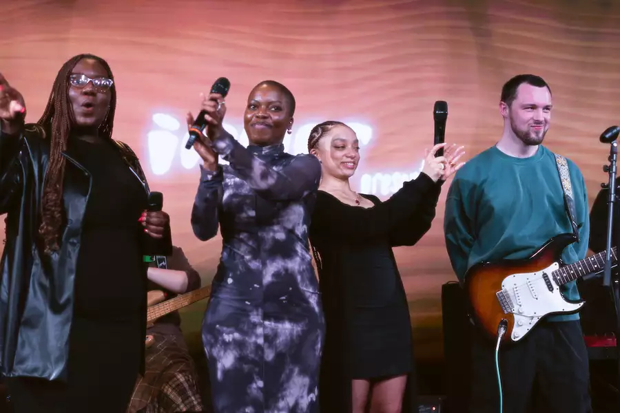 Four performers stand on stage holding microphones, smiling and engaging with the audience, while a guitarist beside them holds his instrument. The background features warm lighting and blurred text.