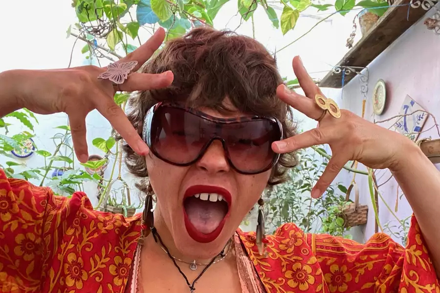 Chia wearing large sunglasses, red lipstick, and fake vampire fangs poses with fingers spread by their face. They have short curly hair, bold rings, and are dressed in a red and orange patterned top, surrounded by plants.