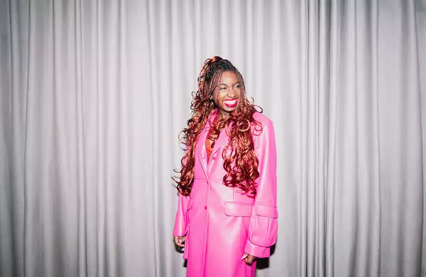 Factory+ Editor in Residence Kemi Alemoru wears a pink trench coat and smiles at the camera