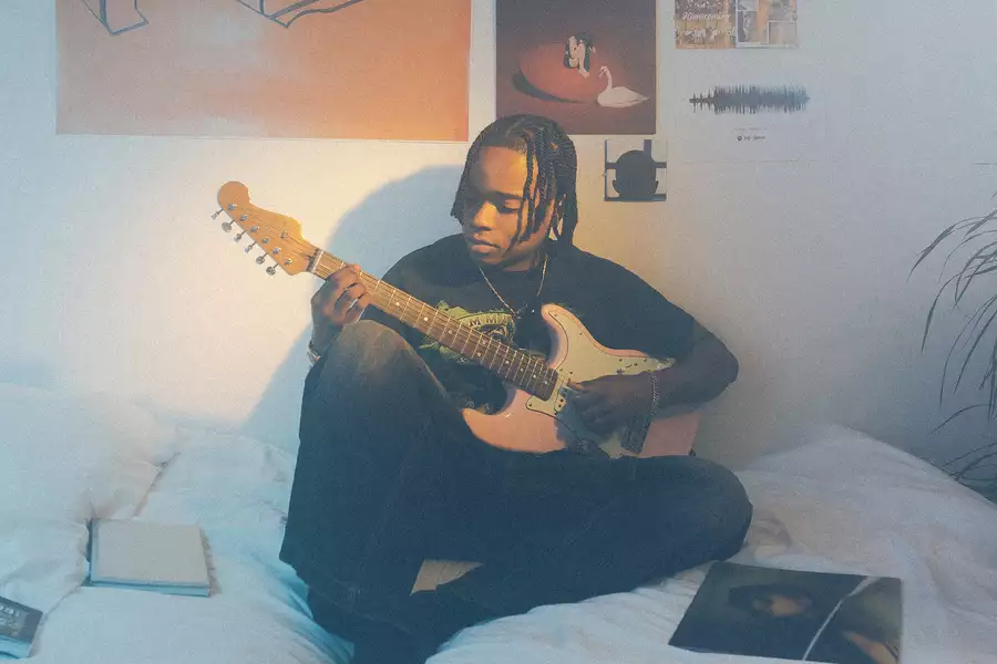 Lei Hope sat on a bed with a white duvet cover playing the electric guitar. Behind them is a wall covered with posters.