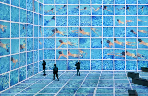 Three people stood in the David Hockney exhibition at Lightroom. His swimming pool paintings are shown on the screens.