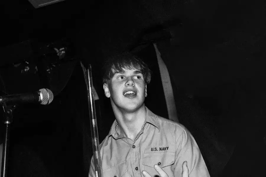 Adam Statham wearing a shirt with U.S. NAVY lettering stands near a microphone on stage, looking slightly upward with his mouth open as if speaking or singing. The background is dark and shadowy.