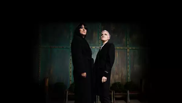 Anna Phoebe and Mary Anne Hobbs dressed in black stand against a rustic metal background, looking upwards.
