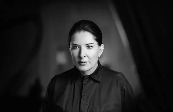 A black-and-white portrait of Marina Abramović, who is shown from the shoulders up, wearing a dark collared shirt. Her hair is neatly pulled back. The background is softly blurred.