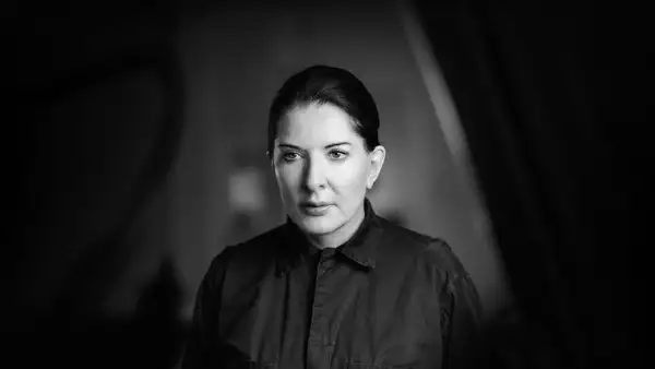 A black-and-white portrait of Marina Abramović, who is shown from the shoulders up, wearing a dark collared shirt. Her hair is neatly pulled back. The background is softly blurred.