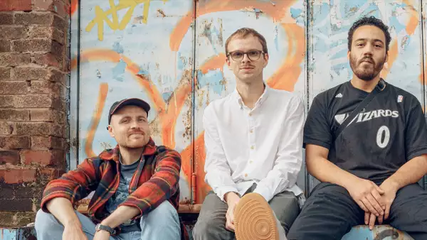 Three members of BRUME sit in front of a wall with colourful graffiti. One wears a red plaid shirt and cap, another wears glasses and a white shirt, and the third wears a black jersey.