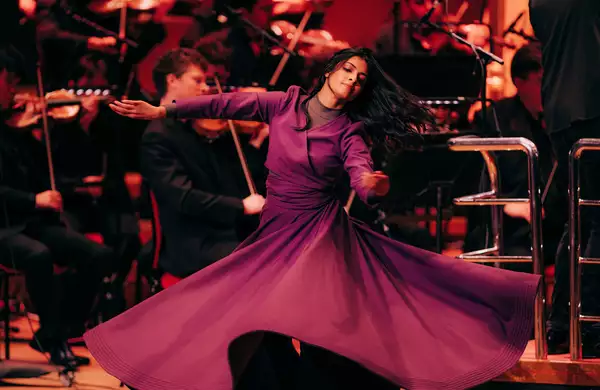 A dancer in a flowing purple dress gracefully spins on stage. Behind her, an orchestra dressed in black plays classical instruments.