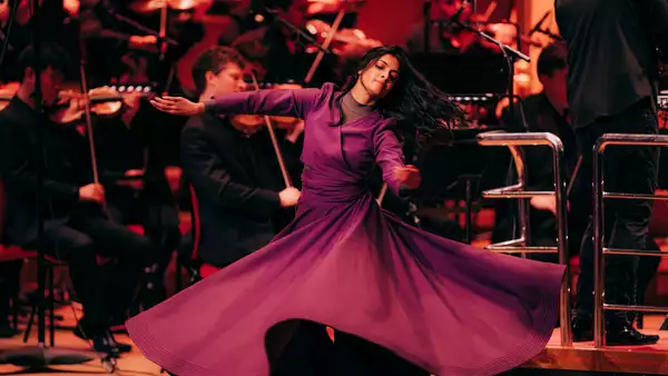 A dancer in a flowing purple dress gracefully spins on stage. Behind her, an orchestra dressed in black plays classical instruments.