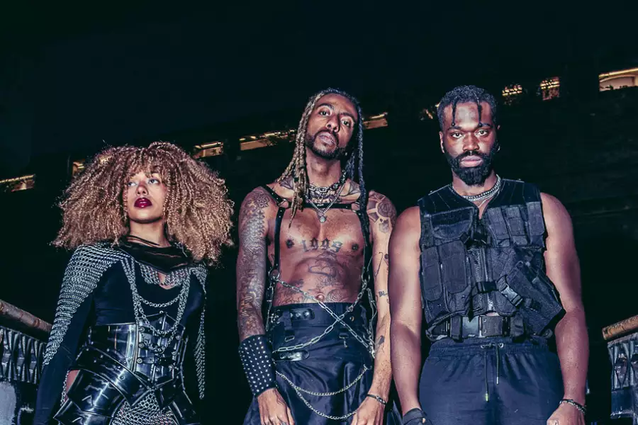 Three members of Third Kulture wearing black outfits and chains. They are stood on a bridge at night.