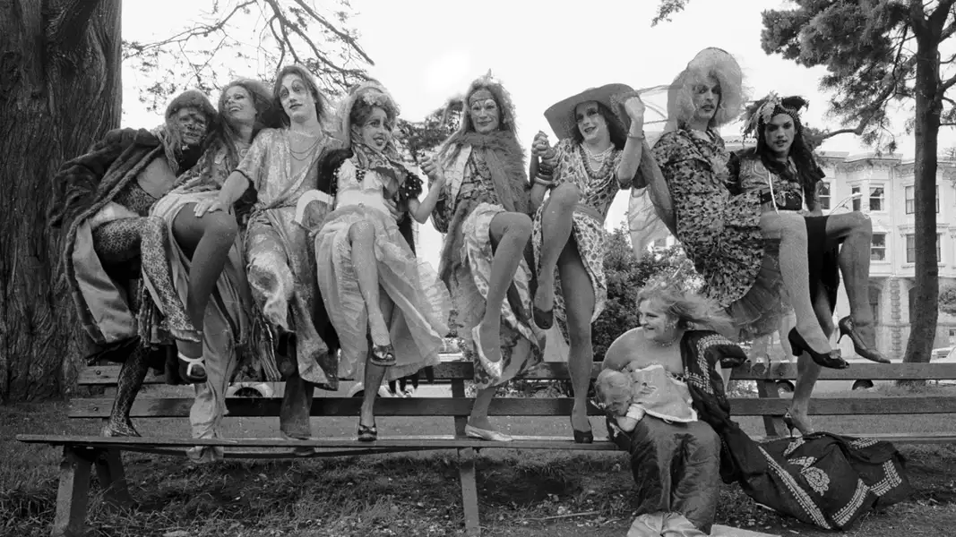 Black and white still from David Weissman's The Cockettes showing a group of people dressed in gowns sat on a fence