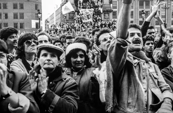 A large crowd of people, bundled in coats, clap, cheer, and hold signs at a protest. The photo is black and white.