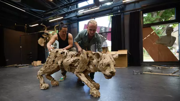 Two people operate a large, detailed animal puppet made from cardboard in a studio space.