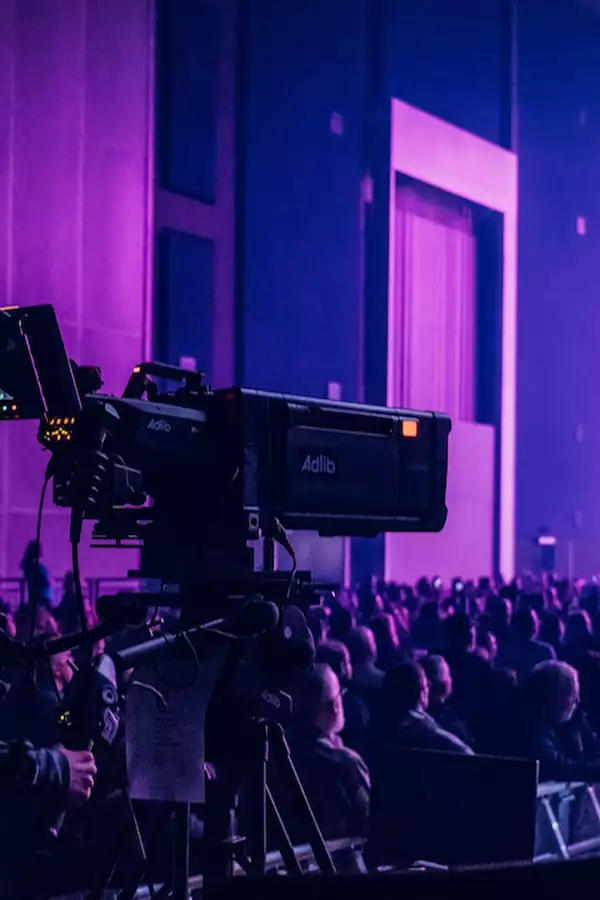A camera operator films a live event in the Warehouse, which is filled with people. There is dramatic purple lighting with multiple cameras and bright stage beams.
