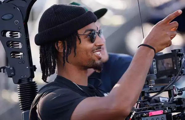 A young Black man wearing sunglasses and a beanie hat operates a video camera.