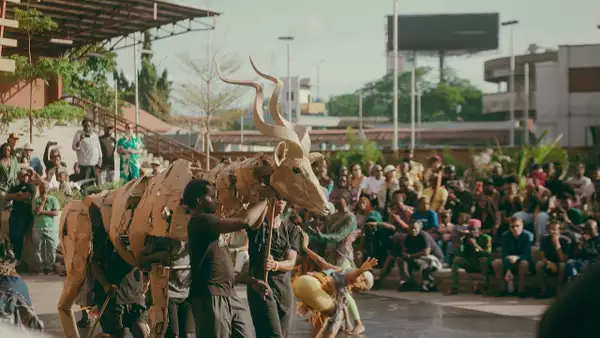 Performers operate a large cardboard antelope puppet in front of a seated outdoor audience.