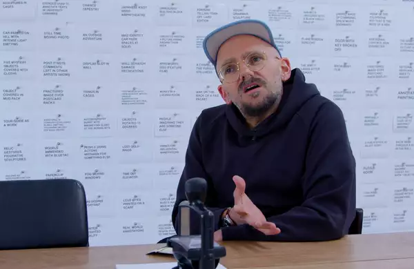 The artist Ryan Gander in his studio. He is a white man, wearing a black hoody and trousers and a multi-coloured cap.