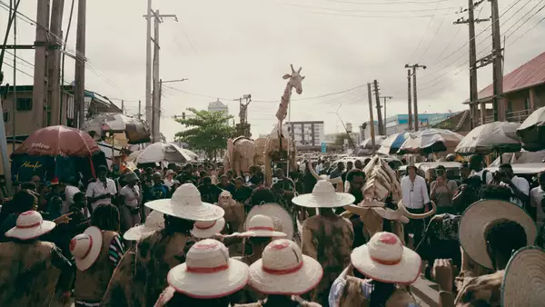 A large crowd of people at an outdoor market wearing hats. They are looking at large giraffe and elephant puppets.