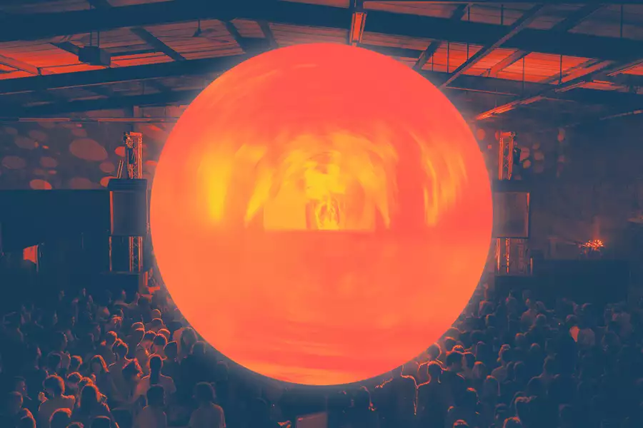A large, glowing orange orb is suspended above a crowd in a dark indoor venue, casting warm light over the audience and the industrial-style ceiling.