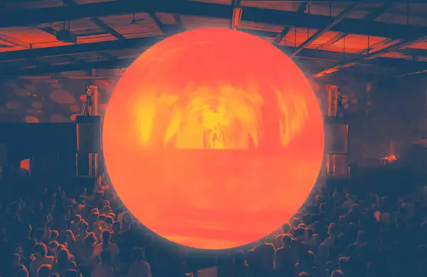 A large, glowing orange orb is suspended above a crowd in a dark indoor venue, casting warm light over the audience and the industrial-style ceiling.
