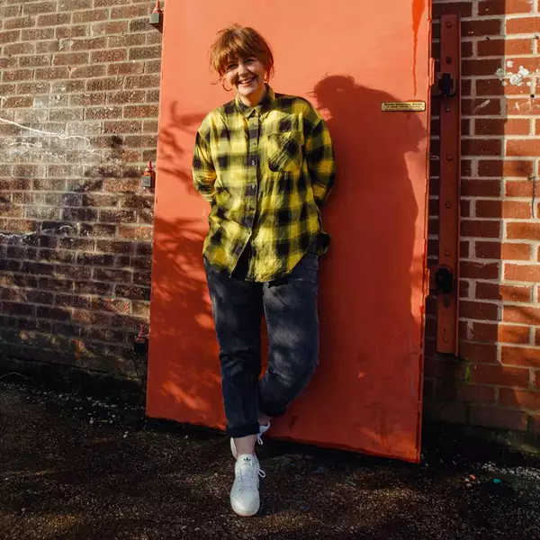 Test Card Girl stood outside against an orange door. She has brown hair in a bun and is wearing a black and yellow check shirt, black jeans and white trainers.