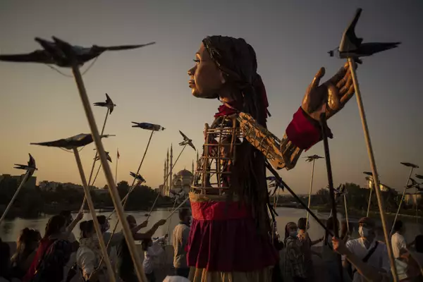 The little Amal puppet stretches her hands wide as wooden birds, held up on sticks by a crowd of people, surround her. In the dusky background is Sabancı Central Mosque in Adana.