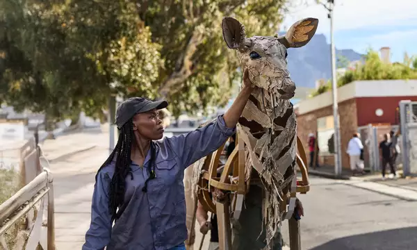 A woman in a cap touches the head of a life-sized giraffe puppet made from cardboard and wood, as part of The Herds. The background shows an urban environment with people watching and a scenic view of trees and mountains.