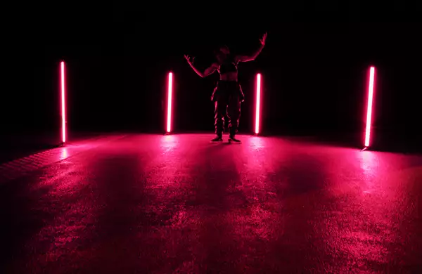 A person stands in a dimly lit space between four vertical neon lights emitting a pink glow, their arms raised in a dramatic pose